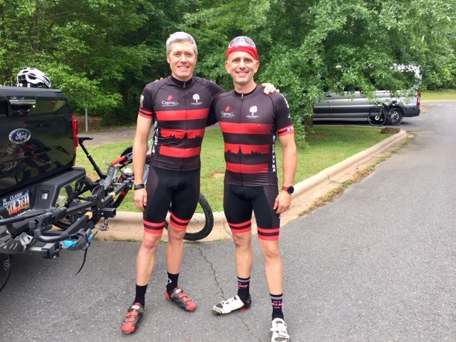 alt=Photo of 2 men wearing cycling jerseys posing together and smiling at the camera