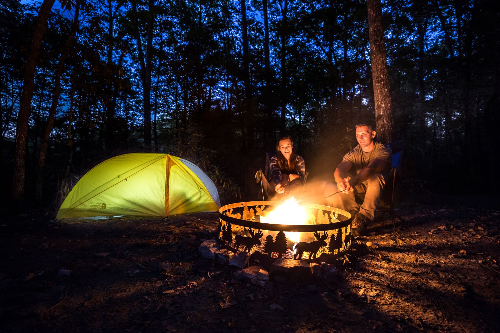 Photo of a couple roasting marshmallows over a campfire next to their tent at the Anne Springs Close Greenway