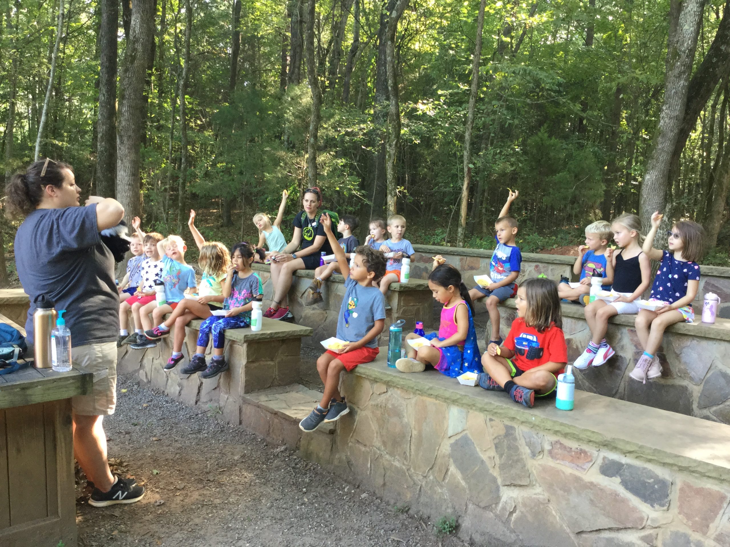 alt=Photo of children raising their hands while sitting outside of the Anne Springs Close Greenway