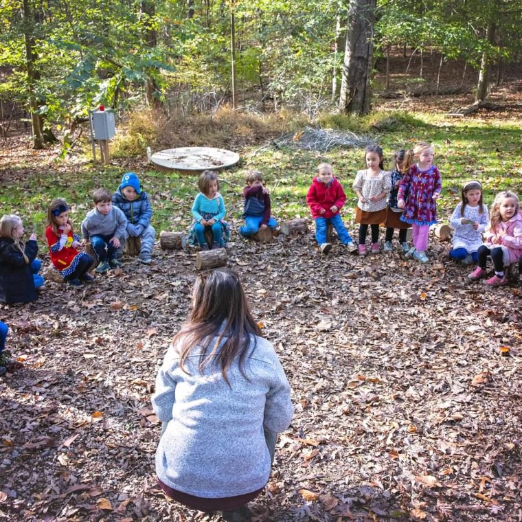 alt=Photo of children sitting on logs in a semi-circle looking at their teacher