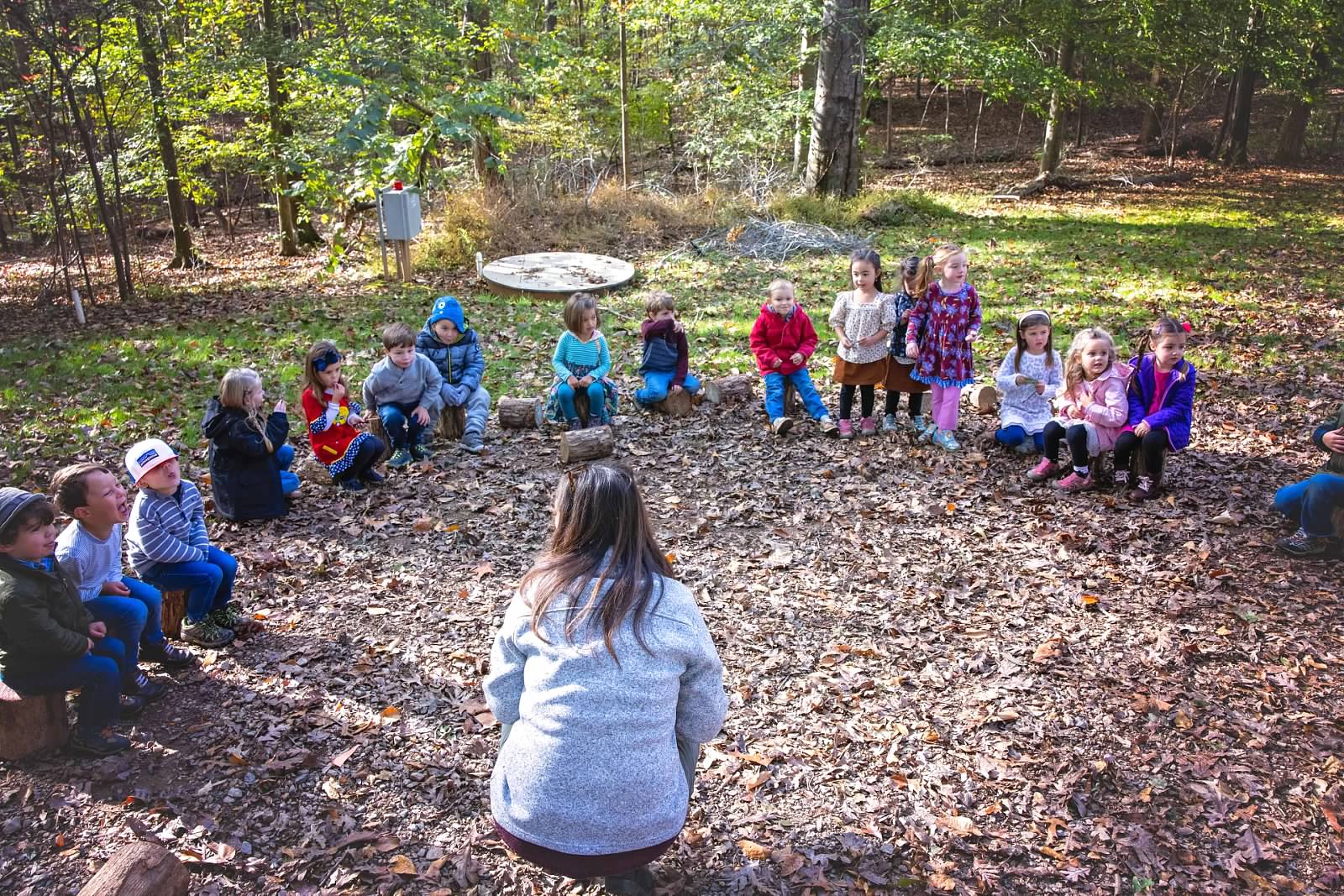 alt=Photo of children sitting on logs in a semi-circle looking at their teacher