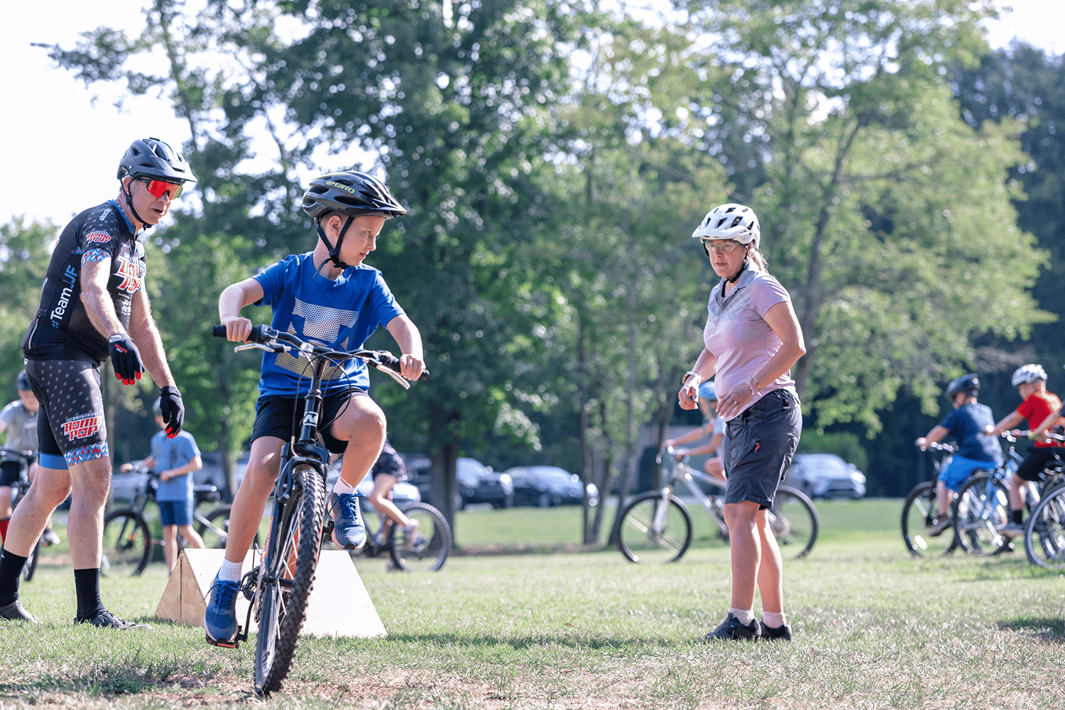 alt=Photo of young boy riding a bike while adult leaders watch him