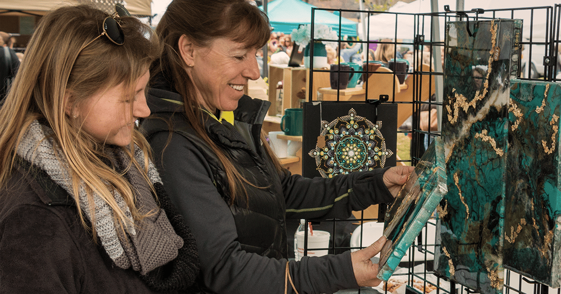 alt=Photo of 2 women smiling at an art piece at the Winter Holiday Artisan Market
