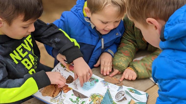 A group of preschool-age children sit at a picnic table outside while reading a book about nature.